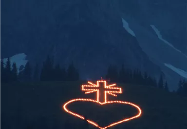 Ein beleuchtetes Herz mit einem Kreuz in den Bergen bei Dämmerung. Die Lichter strahlen in der dunklen Umgebung.