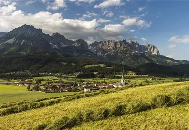 Eine malerische Landschaft mit hohen Bergen im Hintergrund und einem charmanten Dorf im Tal. Die Wiesen sind grün und die Himmel ist teilweise bewölkt.
