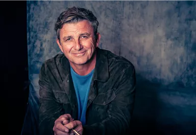 Ein lächelnder Mann mit grau melierten Haaren sitzt vor einem neutralen Hintergrund. Er trägt eine Jacke und ein T-Shirt und schaut freundlich in die Kamera.