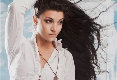 Eine junge Frau mit langen, fließenden Haaren trägt ein weißes Hemd. Sie blickt nachdenklich und hat eine Kette um den Hals.