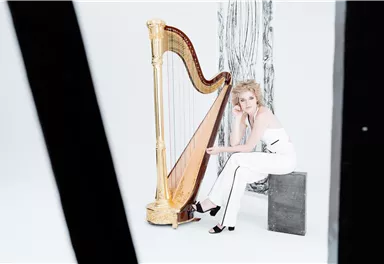 Eine Frau sitzt neben einer goldenen Harfe in einem minimalistischen, hellen Raum. Sie trägt ein elegantes, weißes Outfit und wirkt nachdenklich.