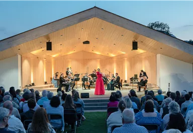 Eine Musikerin in einem roten Kleid steht im Mittelpunkt einer Bühne und singt. Das Publikum sitzt gespannt davor, umgeben von einer schönen Abendkulisse.