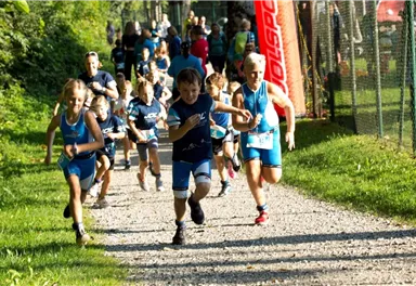 Eine Gruppe von Kindern in Sportkleidung rennt auf einem Kiesweg. Im Hintergrund sind weitere Teilnehmer und eine Flagge zu sehen.