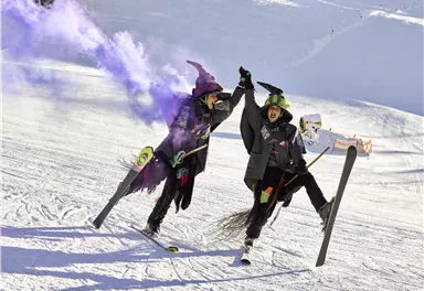 Zwei verkleidete Hexen feiern auf der Skipiste und halten sich an den Händen. Bunte Rauchfackeln umgeben sie in der verschneiten Landschaft.