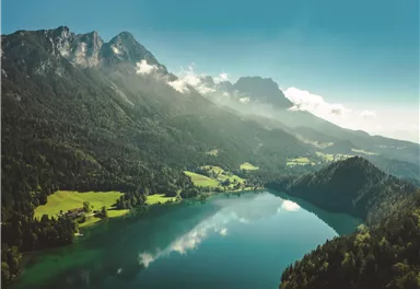 Eine atemberaubende Landschaft mit einem klaren See, umgeben von grünen Wäldern und majestätischen Bergen. Der Himmel ist blau und es gibt einige Wolken in der Ferne.