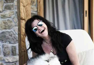 A laughing woman sits on a couch and hugs a dog. The dog enjoys the attention and both appear happy.