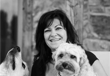 A woman is sitting smiling and holding a small dog in her arms. Next to her stands a wolf, and they both have friendly expressions.