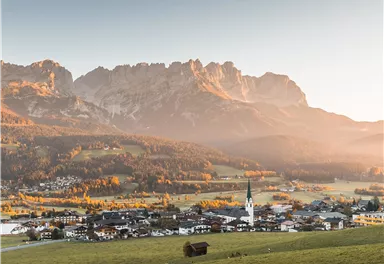 Eine malerische Berglandschaft mit einem kleinen Dorf im Vordergrund. Die Sonne geht über den majestätischen Bergen auf und taucht alles in warmes Licht.