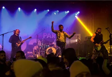 Eine Band spielt live auf der Bühne, während die Menge tanzt und jubelt. Das Licht ist bunt und schafft eine energiegeladene Atmosphäre.