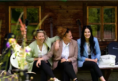 Drei Frauen sitzen lächelnd auf einer Bank vor einem Holzhaus. Im Hintergrund sind Gitarren und Pflanzen zu sehen.