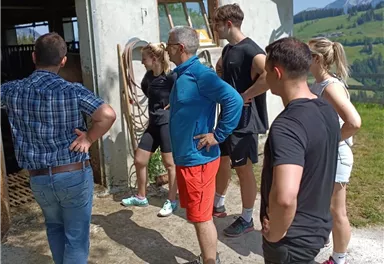 A group of people is standing outside in front of a farmhouse. They are looking at something curiously and seem to be interested.