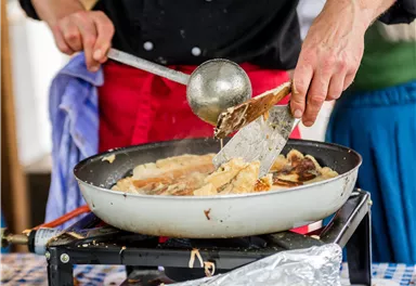 Eine Person kocht in einer Pfanne auf einem Gasherd. Man sieht, wie sie mit einem Spatel das Essen wendet.