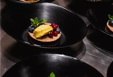 Eine elegante Dessertpräsentation in schwarzen Schalen. Die Desserts sind mit Beeren und einer gelben Sorbetkugel dekoriert.