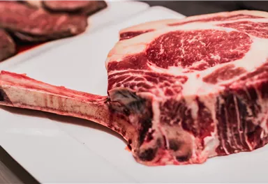 Ein großes, rohes Tomahawk-Steak auf einem weißen Teller. Das Fleisch hat eine ausgeprägte Marmorierung und einen langen Knochenschaft.