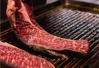 Ein Koch hebt ein Stück rotes Fleisch auf einen Grill. Die Fleischstücke zeigen eine schöne Marmorierung.