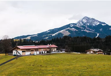 Ein malerisches Landschaftsbild mit einem Bauernhaus im Vordergrund und einem majestätischen Berg im Hintergrund. Der Himmel ist bewölkt und die Wiesen sind grün.