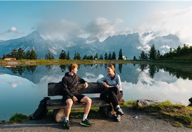 Zwei Personen sitzen auf einer Bank am Ufer eines Sees. Im Hintergrund sind Berge und ein klarer Himmel zu sehen.