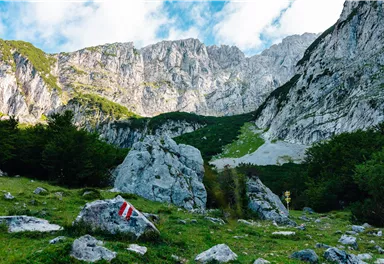 Eine beeindruckende Berglandschaft mit hohen Felsen und einer grünen Wiese. Am Hang sind Wanderzeichen sichtbar.