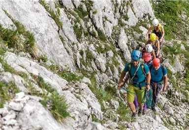 Eine Gruppe von Bergsteigern erklimmt einen felsigen Pfad in den Bergen. Sie tragen Helme und Kletterausrüstung.
