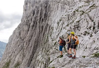 Drei Kletterer stehen an einer steilen Felswand und betrachten die Landschaft. Sie tragen Helme und Kletterausrüstung.