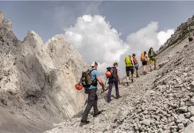Eine Gruppe von Menschen wandert auf einem steilen, steinigen Weg in den Bergen. Im Hintergrund sind hohe Felsen und ein bewölkter Himmel zu sehen.