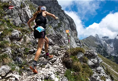 Eine Person läuft auf einem steinigen Pfad in den Bergen. Die Landschaft ist beeindruckend mit grünen Wiesen und steilen Felsen.