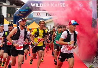 Läufer überschreiten die Ziellinie bei einem Wettkampf im Freien. Im Hintergrund ist bunte Rauchfontäne und eine Zielmarke zu sehen.