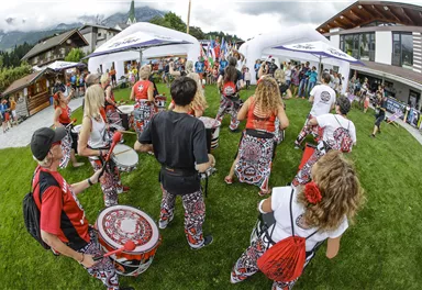 Eine fröhliche Menschenmenge nimmt an einem Fest teil. Im Vordergrund spielen Musiker mit Trommeln und bunte Tänzerinnen und Tänzer sind zu sehen.