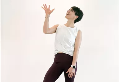 Eine Person steht auf einer Yogamatte und jongliert mit einem Ball. Sie trägt ein sportliches Outfit und lächelt dabei.