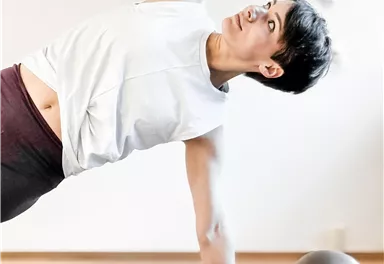 Eine sportliche Frau führt eine Seitstütz-Übung auf einer Yogamatte aus. Neben ihr liegt eine Fitnesskugel.