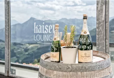 Eine rustikale Barrique-Fass mit zwei Champagnerflaschen und einer kleinen Pflanze. Im Hintergrund sind Berge und eine Fensterbeschriftung zu sehen.