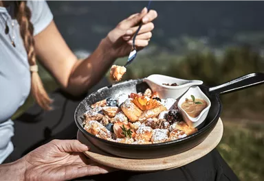 A narrow gravel path winds through an autumn forest. Colorful leaves and tall trees surround the landscape, while mountains can be seen in the background.