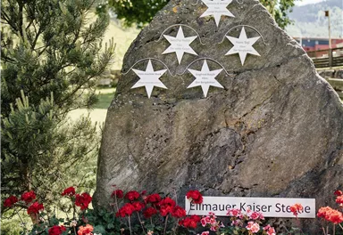 A large rock with white stars is surrounded by colorful flowers. The inscription on the rock reads "Elmauer Kaiser Straße".