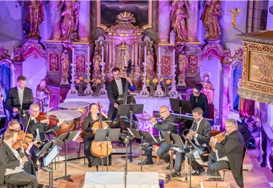 Ein Orchester spielt in einer festlich dekorierten Kirche. Die Musiker sind auf der Bühne und die Umgebung ist mit bunten Lichtern beleuchtet.