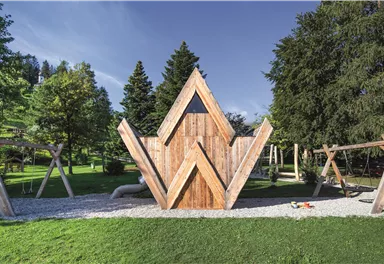 A modern wooden playground with a striking triangular playhouse. In the background, trees and swings can be seen.
