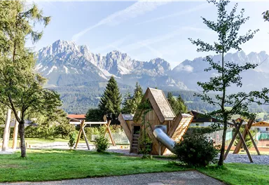 A beautiful playground in a park with a unique play equipment and shade-giving trees. In the background, impressive mountains are visible.
