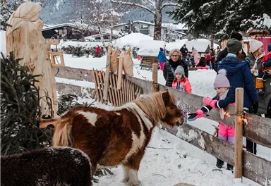 Ein Weihnachtsmarkt mit Schnee, Tieren und fröhlichen Menschen. Im Hintergrund sind schneebedeckte Berge und eine festliche Atmosphäre zu sehen.