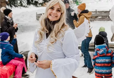 Eine fröhliche Person in einem weißen Engel-Kostüm steht in einer verschneiten Landschaft. Im Hintergrund sind Kinder und Erwachsene, die sich in einem winterlichen Festumfeld versammeln.