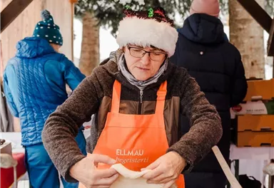 Eine Person in einer Weihnachtsmütze steht an einem Stands mit einem Frittiergerät und bereitet Speisen zu. Im Hintergrund sind weitere Personen zu sehen, die sich in warmer Kleidung aufhalten.