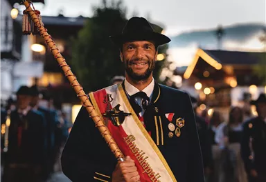 Ein Mann in traditioneller Kleidung hält eine Fahne und lächelt stolz. Im Hintergrund sind weitere Personen in Trachten zu sehen, die an einem festlichen Umzug teilnehmen.