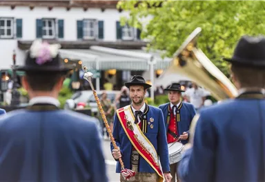 Eine traditionelle Trachtenparade mit Menschen in blauen Jacken und Hüten. Im Vordergrund steht ein Mann mit einem Zepter und einer Ehrenbinde.