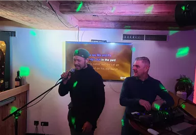 Zwei Männer stehen in einer Bar. Einer singt mit einem Mikrofon, während der andere am Mischpult arbeitet.