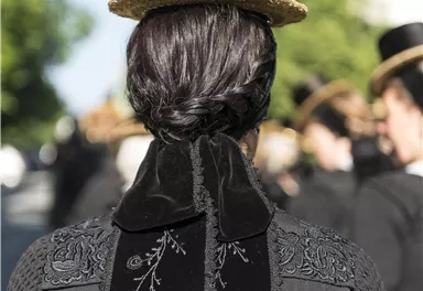 Eine Person mit einem traditionellen schwarzen Kleid und einem eleganten Hut steht im Vordergrund. Im Hintergrund sind weitere Personen in ähnlicher Kleidung zu sehen.