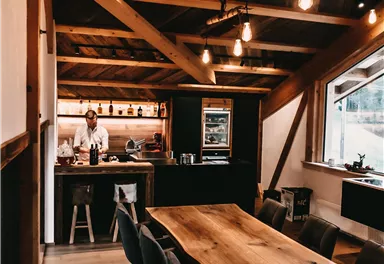 Ein modernes Esszimmer mit einem großen Holztisch und stilvollen Stühlen. Im Hintergrund ist eine offene Küche mit einem Koch zu sehen.