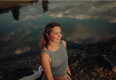 Eine Frau sitzt am Ufer eines Sees und schaut nachdenklich ins Wasser. Die Umgebung spiegelt sich im ruhigen Wasser und schafft eine friedliche Atmosphäre.