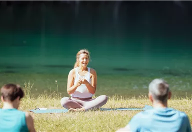 Eine Gruppe von Menschen macht Yoga am Ufer eines ruhigen Sees. Eine Frau sitzt lächelnd im Vordergrund und leitet die Übung.