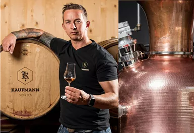 Ein Fachmann steht neben Weinlagertonnen und hält ein Glas in der Hand. Im Hintergrund sind Kupfergeräte zur Destillation zu sehen.