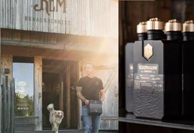 Ein Mann trägt eine Flasche aus schwarzem Glas und verlässt ein Holzgebäude mit der Aufschrift „Mohlberg Rem“. Ein Hund läuft neben ihm.