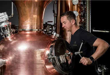 Ein Mann arbeitet an einer kupfernen Destillieranlage. Im Hintergrund sind Druckmessgeräte sichtbar.