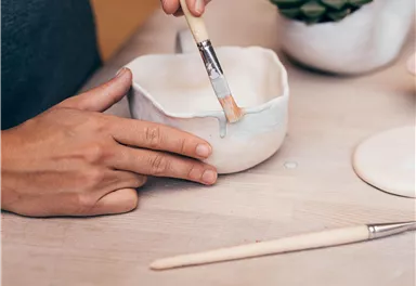 A person is painting with a brush on a ceramic bowl. In the background, additional decorative objects can be seen.